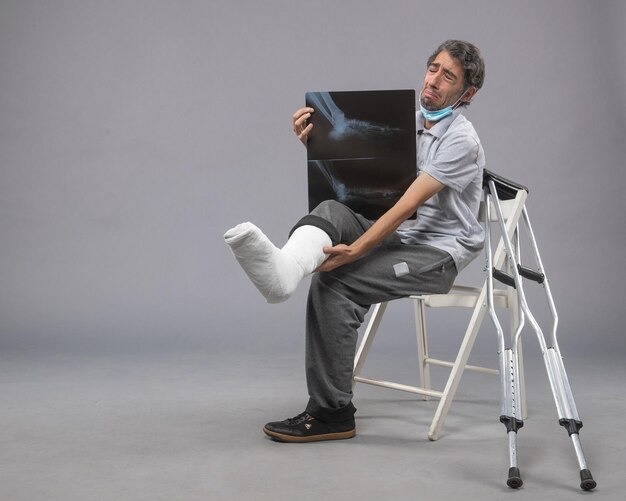 Vooraanzicht jonge man zit met gebroken voet en houdt röntgenfoto ervan op grijze muur pijn been gebroken ongeval voet twist man