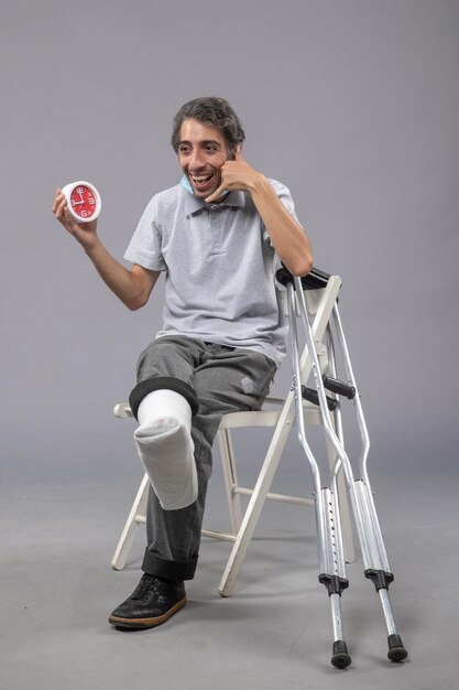 Vooraanzicht jonge man zit met gebroken voet vastgebonden verband en houdt klok vast op grijze vloer voet draai mannelijk ongeval pijn been