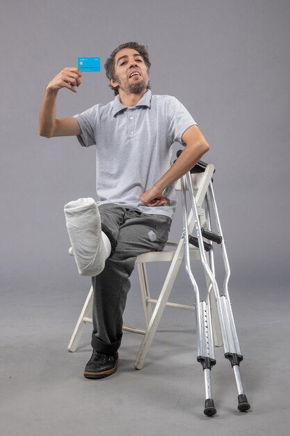 Vooraanzicht jonge man zittend met gebroken voet met blauwe bankkaart op de grijze muur gebroken ongeval voet pijn twist man