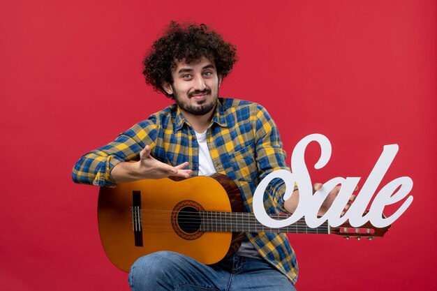 Vooraanzicht jonge man zittend met gitaar op de rode muur verkoop speel concertmuziek kleur applaus live muzikant