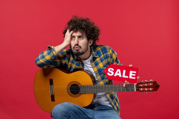 Vooraanzicht jonge man zittend met gitaar op rode muur concert live muziek kleur muzikant verkoop spelen