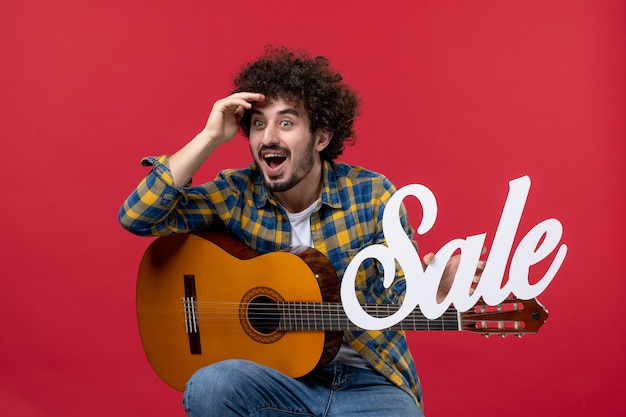 Vooraanzicht jonge man zittend met gitaar op rode muur kleur applaus muzikant spelen band concert live verkoop