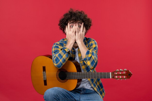 Vooraanzicht jonge man zittend met gitaar op rode muur live concert muziek kleur muzikant applaus band spelen