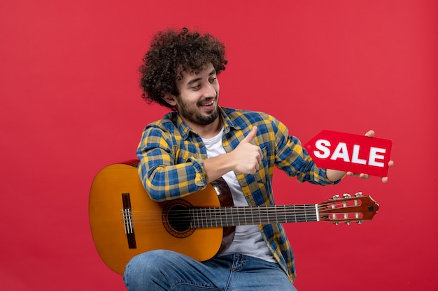 Vooraanzicht jonge man zittend met gitaar op rode muur live muziek kleur applaus muzikant verkoop spelen