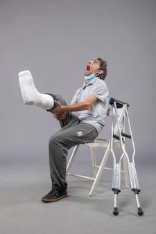 Vooraanzicht jonge man zittend met verbonden gebroken voet schreeuwend van pijn op de grijze muur pijn been ongeval draai mannelijke voet