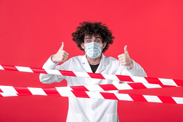 Vooraanzicht jonge mannelijke arts in medisch uniform en masker op de rode achtergrond isolatie ziekenhuis covid-gezondheid quarantaine gevaar waarschuwingsstrip