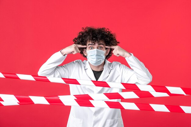 Vooraanzicht jonge mannelijke arts in medisch uniform en masker op rode achtergrond gevaar covid-gezondheid quarantaine waarschuwing ziekenhuis