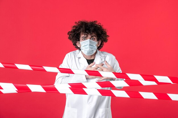 Vooraanzicht jonge mannelijke arts in medisch uniform en masker op rode achtergrond gevaar isolatie ziekenhuis covid-gezondheid quarantaine waarschuwingsstrip