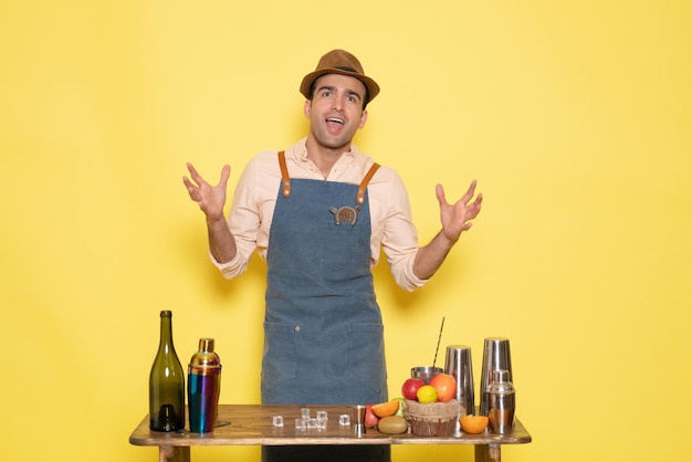 Vooraanzicht jonge mannelijke barman voor tafel met shakers drankjes op gele achtergrond