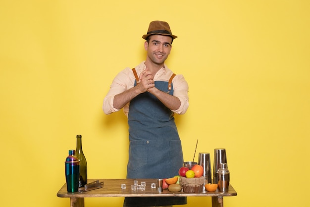 Gratis foto vooraanzicht jonge mannelijke barman voor tafel met shakers drankjes op gele achtergrond