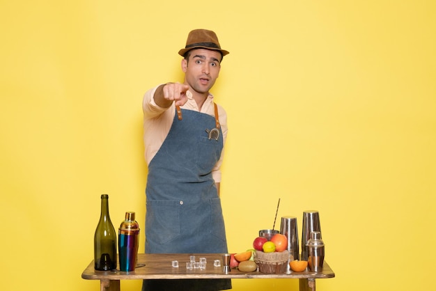 Vooraanzicht jonge mannelijke barman voor tafel met shakers drankjes op gele achtergrond