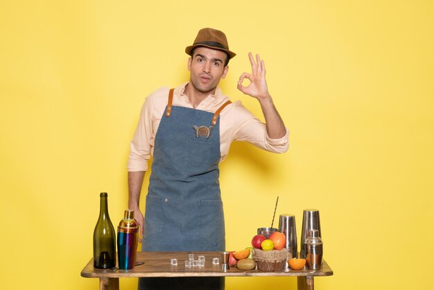 Vooraanzicht jonge mannelijke barman voor tafel met shakers en drankjes poseren op gele achtergrond