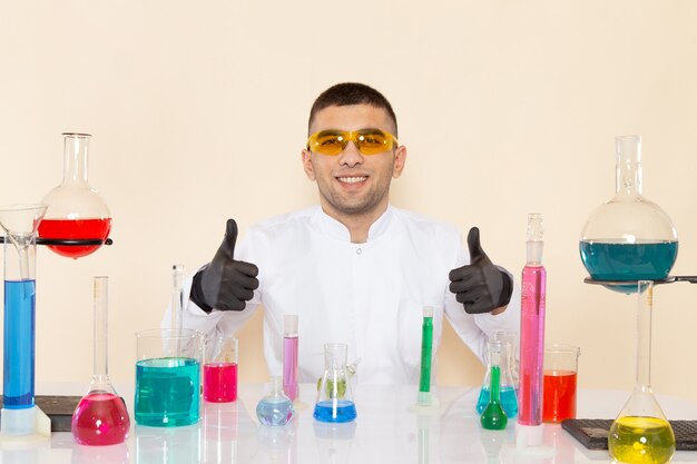 Vooraanzicht jonge mannelijke chemicus in wit speciaal pak zit tafel met oplossingen en lachend op crème muur lab chemie wetenschappelijk experiment