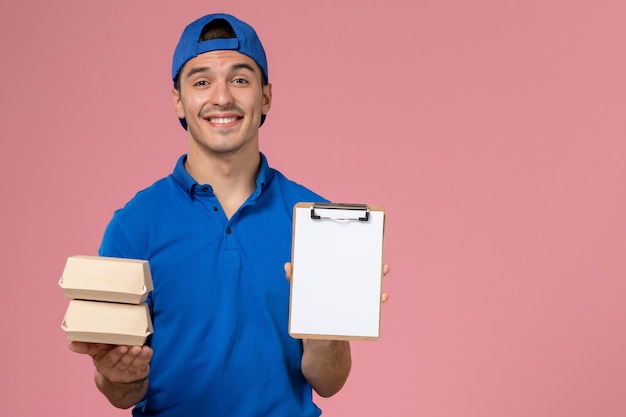 Vooraanzicht jonge mannelijke koerier in blauwe uniforme cape die kleine pakketjes van het leveringsvoedsel met blocnote houdt en op lichtroze muur glimlacht