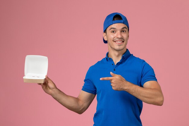 Vooraanzicht jonge mannelijke koerier in blauwe uniforme cape die weinig leeg leveringsvoedselpakket op de roze muur houdt