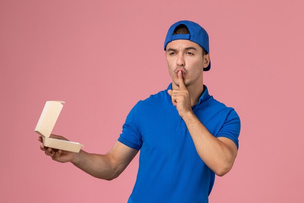 Vooraanzicht jonge mannelijke koerier in blauwe uniforme cape die weinig leeg leveringsvoedselpakket op de roze muur houdt