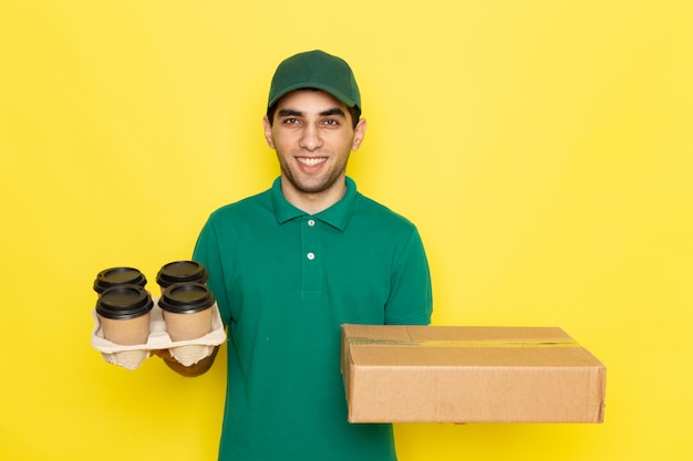 Vooraanzicht jonge mannelijke koerier in groen shirt groen glb bedrijf leveringsdoos en koffiekopjes op geel