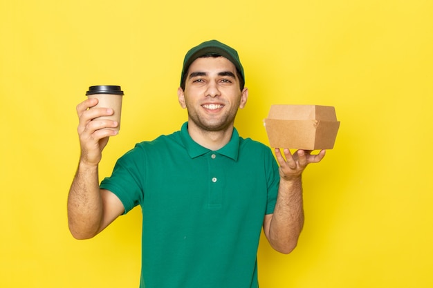 Vooraanzicht jonge mannelijke koerier in groen shirt groen GLB bedrijf leveringspakket en koffiekopje met glimlach op geel