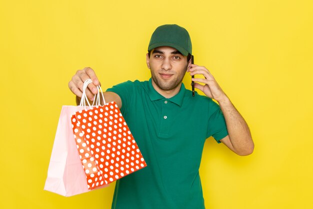 Vooraanzicht jonge mannelijke koerier in groen shirt groene pet praten aan de telefoon en winkelen pakketten op de gele achtergrond service kleur leveren