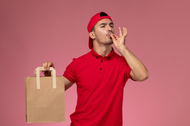 Vooraanzicht jonge mannelijke koerier in rode uniforme cape met papier voedselpakket op roze achtergrond.