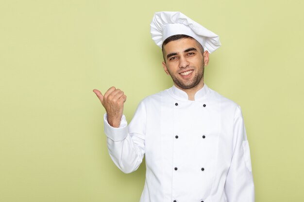 Vooraanzicht jonge mannelijke kok in wit kokkostuum die en vingerteken op groen glimlachen tonen