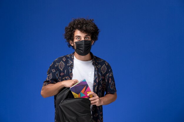 Vooraanzicht jonge mannelijke student die zwart masker draagt en rugzak op lichtblauwe achtergrond houdt.