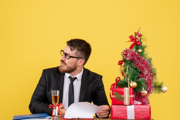 Vooraanzicht jonge mannelijke werknemer achter zijn tafel met cadeautjes en kerstboom op geel