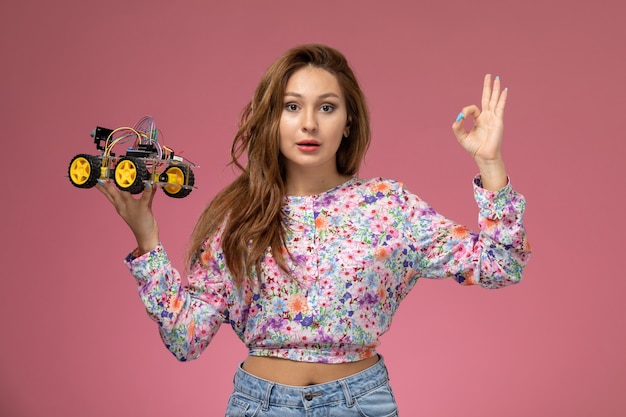 Vooraanzicht jonge mooie vrouw in bloem ontworpen shirt en spijkerbroek houden speelgoedauto poseren op de roze achtergrond