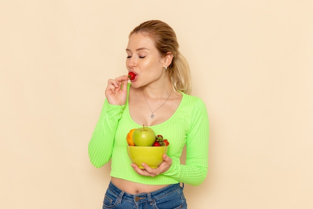 Vooraanzicht jonge mooie vrouw in groen shirt met bord met fruit aardbei eten op de lichte crème muur fruit model vrouw pose