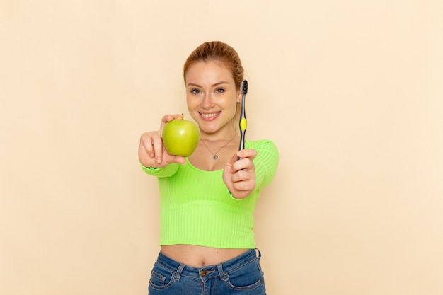 Gratis foto vooraanzicht jonge mooie vrouw in groen shirt met groene appel en tandenborstel op de crème muur fruit model mellow vrouw