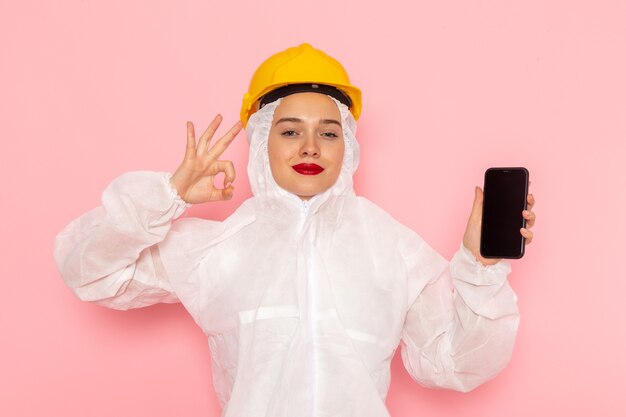 Vooraanzicht jonge mooie vrouw in speciaal wit pak dragen van beschermende helm met telefoon met glimlach op de roze ruimte speciaal pak vrouw