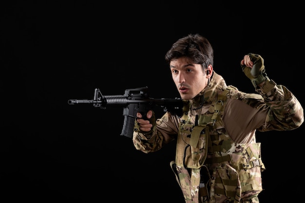 Vooraanzicht jonge soldaat in uniform die zijn geweer op zwarte muur richt