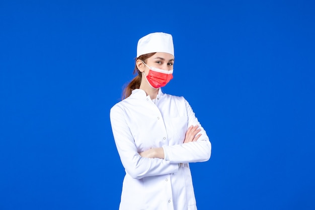 Vooraanzicht jonge verpleegster in medisch pak met rood beschermend masker op blauwe muur