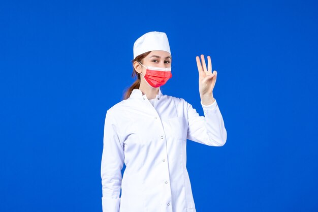 Vooraanzicht jonge verpleegster in medisch pak met rood beschermend masker op blauwe muur