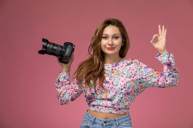 Vooraanzicht jonge vrouw in bloem ontworpen shirt en spijkerbroek met camera glimlachend lichtjes op roze achtergrond