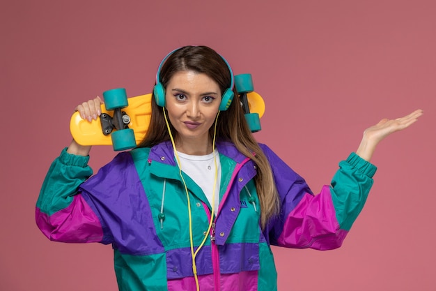 Vooraanzicht jonge vrouw in kleurrijke jas met skateboard op lichtroze muur, model vrouw pose