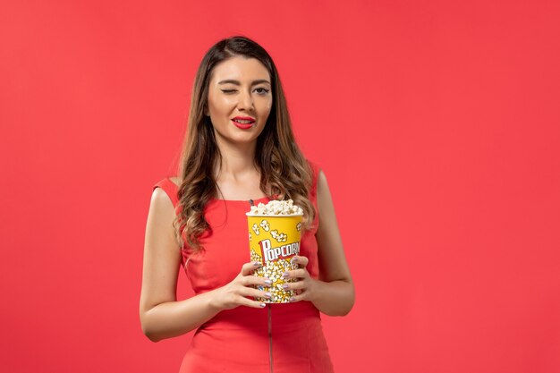 Vooraanzicht jonge vrouw in rood shirt met popcorn kijken naar film knipogen op rode ondergrond