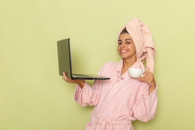 Vooraanzicht jonge vrouw in roze badjas met behulp van laptop en koffie drinken op een lichtgroen oppervlak