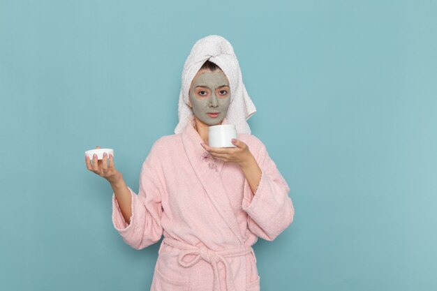 Vooraanzicht jonge vrouw in roze badjas met masker op haar gezicht met crème op het blauwe bureau douche schoonmakende schoonheid zelfzorgcrème