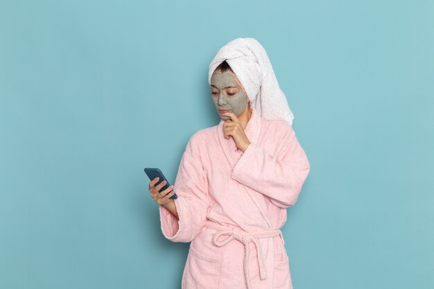 Vooraanzicht jonge vrouw in roze badjas na douche met behulp van haar telefoon op lichtblauwe muur schoonheid water zelfzorg douche schoon