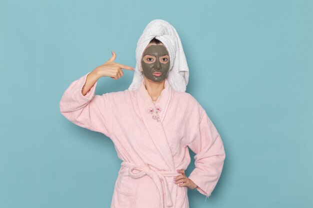 Vooraanzicht jonge vrouw in roze badjas na douche met donker masker op haar gezicht op blauwe muur schoonheid water crème zelfzorg douche badkamer