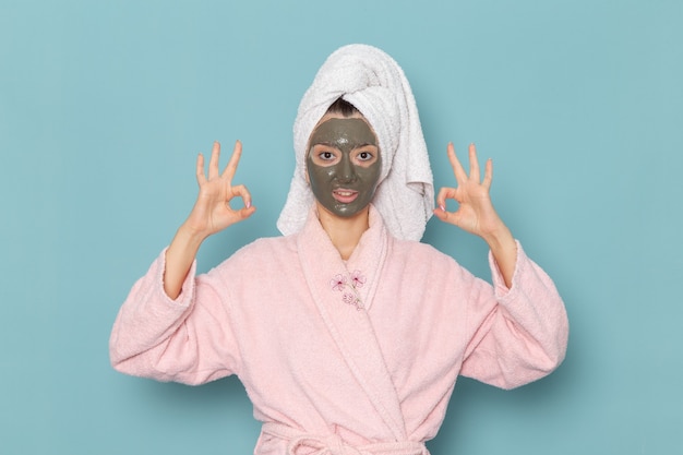 Vooraanzicht jonge vrouw in roze badjas poseren met masker op haar gezicht op de blauwe muur schoonheid water bad crème zelfzorg douche