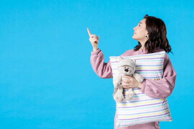 Vooraanzicht jonge vrouw in roze pyjama met kleine speelgoedbeer en kussen op blauwe vrouw