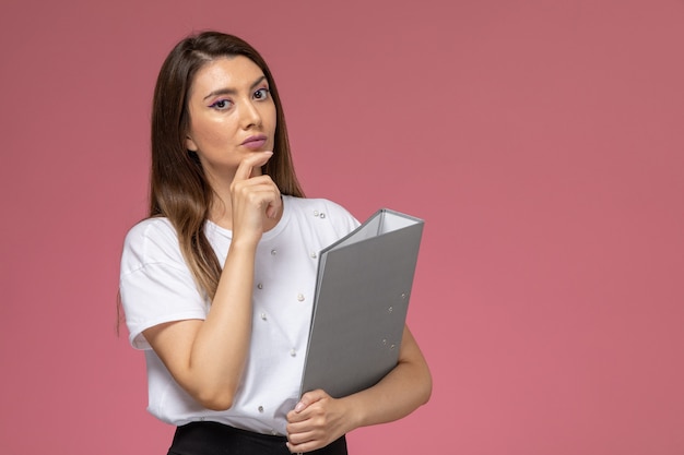 Vooraanzicht jonge vrouw in wit overhemd denken en houden grijs bestand op lichtroze muur, model vrouw pose