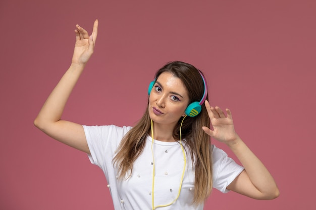 Vooraanzicht jonge vrouw in wit overhemd luisteren naar muziek via gekleurde oortelefoons dansen op roze muur, kleur vrouw pose model