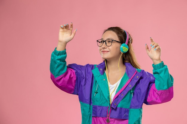 Vooraanzicht jonge vrouw in wit t-shirt ed jas luisteren naar muziek via koptelefoon op de roze achtergrond