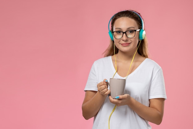 Vooraanzicht jonge vrouw in wit t-shirt gewoon luisteren naar muziek via oortelefoons en kopje met thee op de roze achtergrond te houden