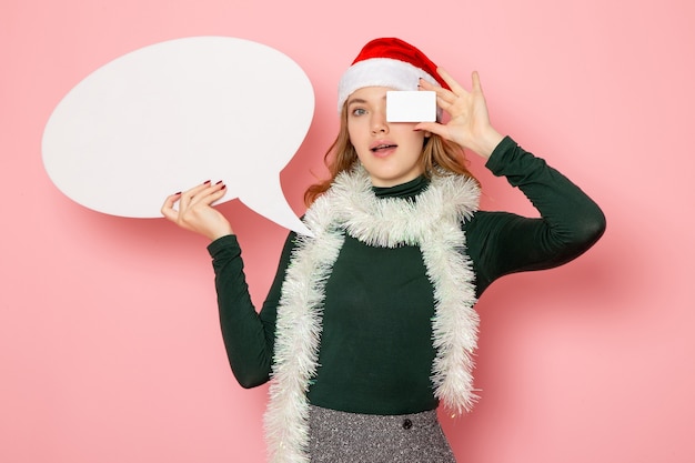 Vooraanzicht jonge vrouw met groot wit bord en bankkaart op roze muur vakantie kerst