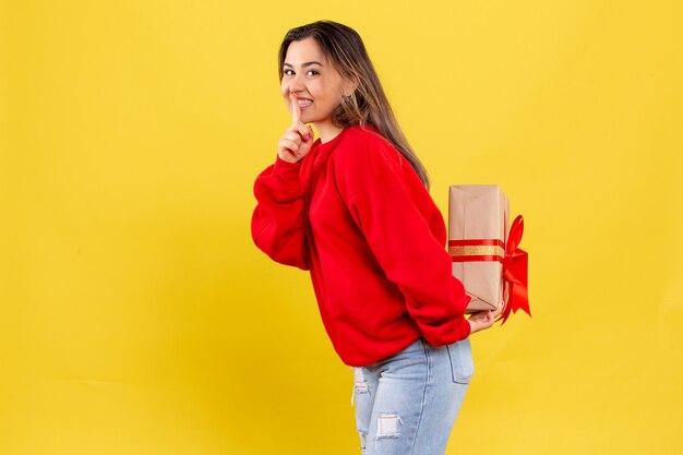 Vooraanzicht jonge vrouw verbergt kerstcadeau achter haar rug op gele achtergrond