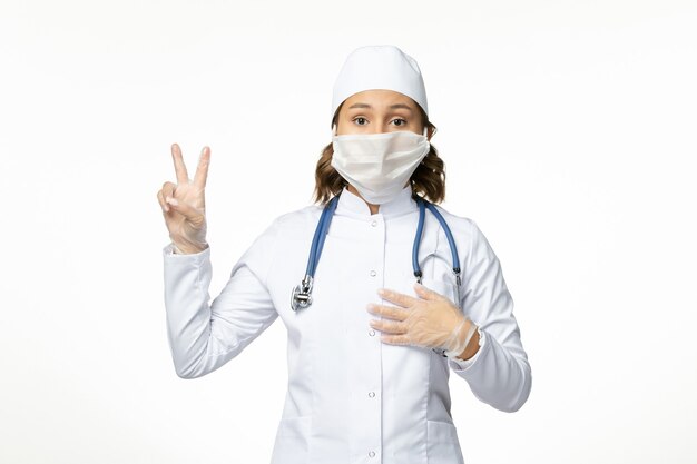 Vooraanzicht jonge vrouwelijke arts die een beschermend steriel masker draagt vanwege het coronavirus op een wit oppervlak